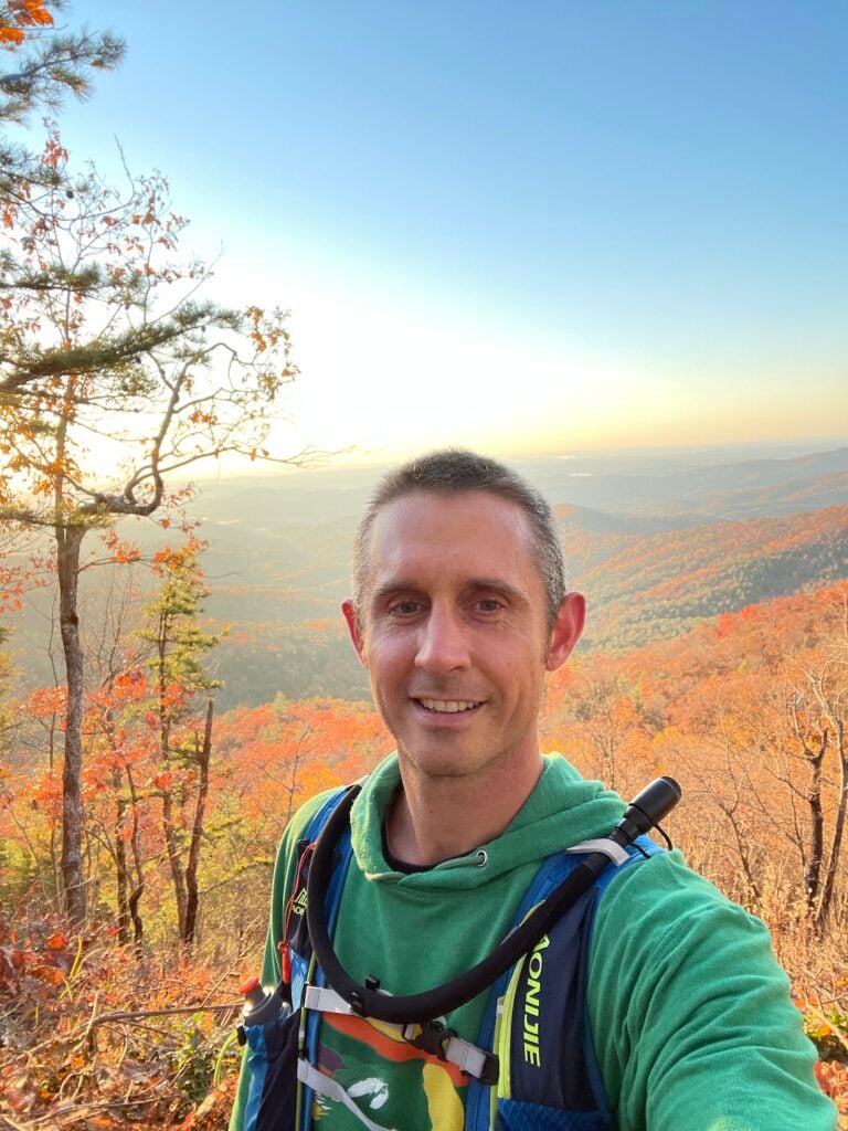 Smiling selfie with many miles of mountains covered in fall leaves in the background.