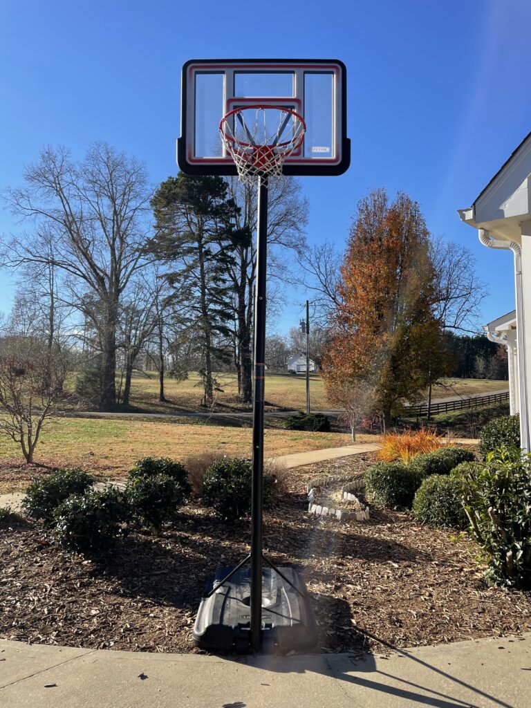 A basketball goal standing at the edge of a driveway with a front yard in the background.