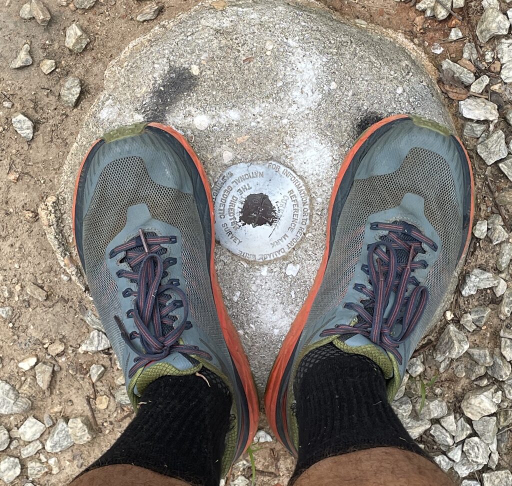 Running shoes positioned next to a metal circle indicating a marker set by the US Geological Survey to mark the summit of Curahee Mountain.