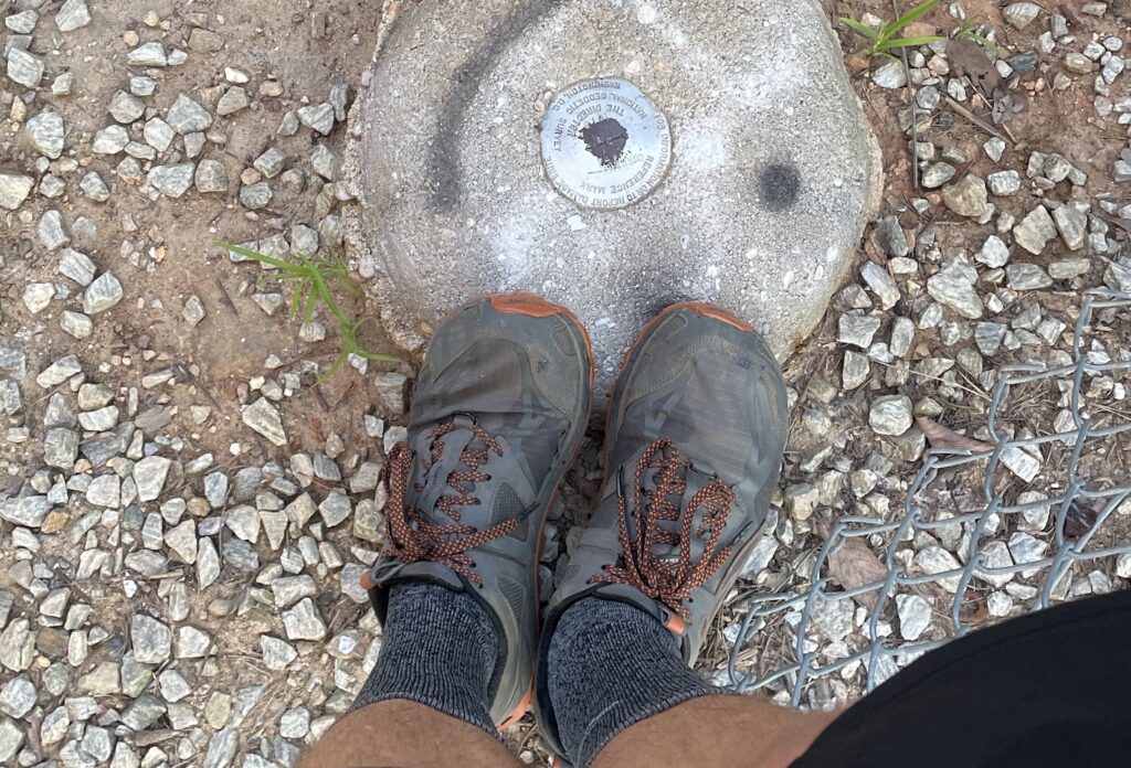 Running shoes positioned next to a metal circle indicating a marker set by the US Geological Survey to mark the summit of Curahee Mountain.