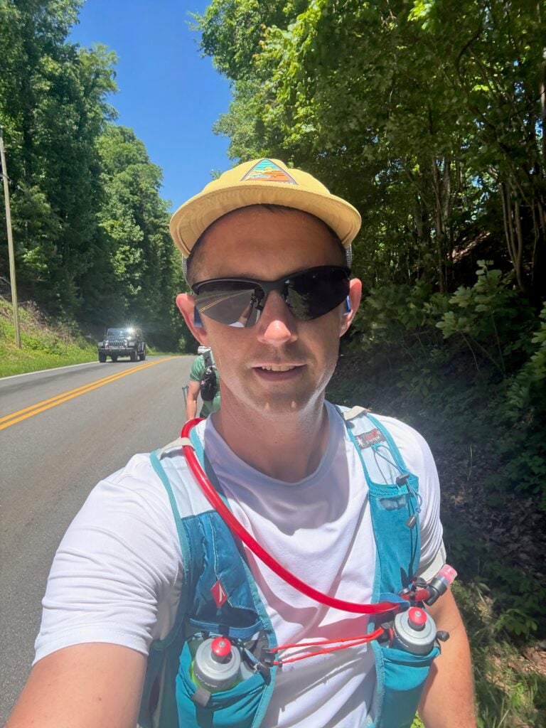 A man in a yellow hat and blue hydration vest takes a selfie while running on a paved road lined with trees